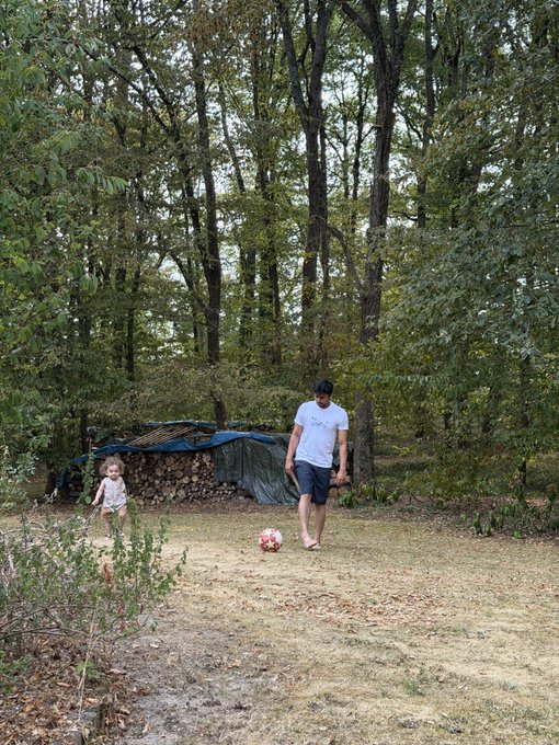 A man wearing a cap, white t-shirt, and dark shorts plays barefoot soccer on a grassy clearing with a young child in the woods.
