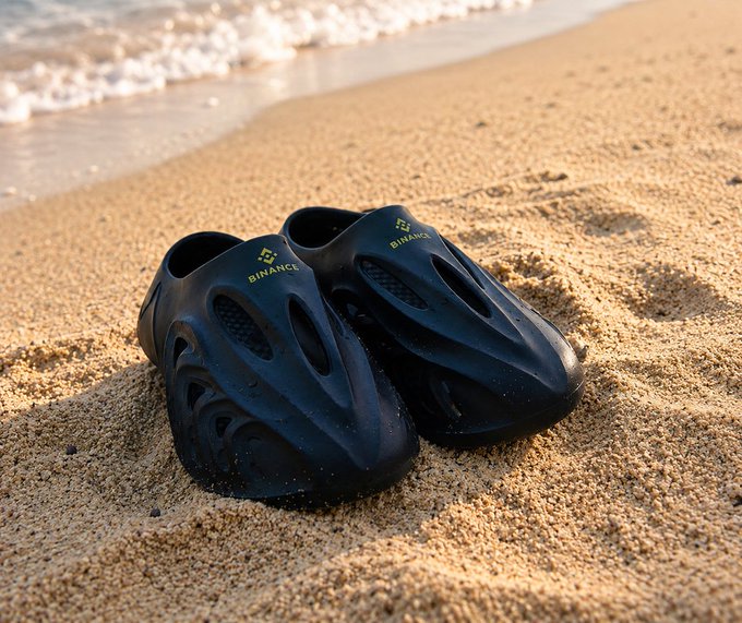 A pair of black slip-on shoes with the Binance logo resting on a sandy beach near the water's edge.