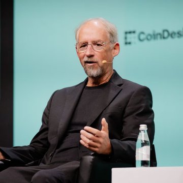An older man with glasses and a gray beard speaks at a CoinDesk event, wearing a dark blazer over a black t-shirt.
