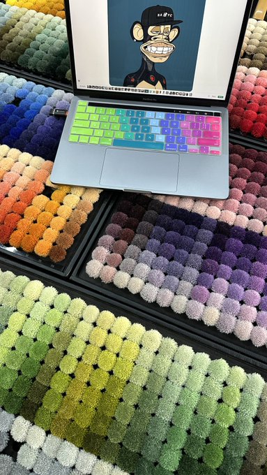 A MacBook laptop with a rainbow-colored keyboard cover sits on a display of multicolored fuzzy keycap samples, with a cartoon character on the screen and Japanese text visible at the bottom.