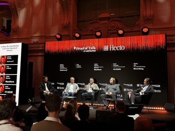 The image shows a group of five people sitting around a table, engaged in a discussion or presentation. They are all dressed professionally, with one person wearing a tie. The setting appears to be an event where they are discussing tokenised markets, as indicated by the text on the screen behind them. The audience is also present, watching and listening attentively to the conversation taking place at the main stage of the event.