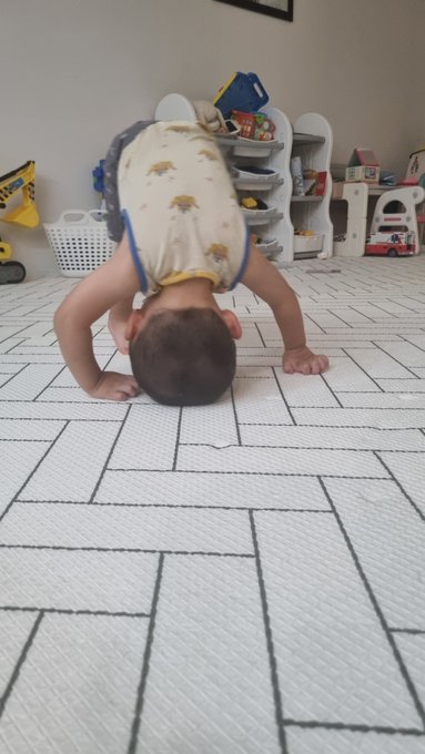 A baby wearing a yellow shirt performing a headstand on a white interlocking floor mat.