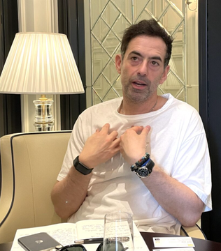 The image shows a man sitting at a table with a laptop, glasses, and a cell phone. The man appears to be wearing a white shirt and has a serious expression as he looks directly into the camera. There are also two cups on the table in front of him. The background features a large window that allows natural light to enter the room, creating an atmosphere for work or study.