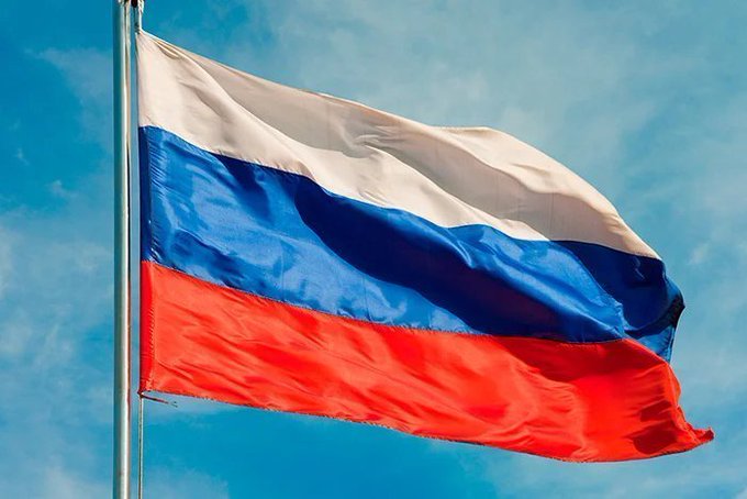 The image shows the Russian flag waving against a blue sky, featuring its horizontal tricolor of white, blue, and red stripes. It visually accompanies the breaking news about Russia's State Duma passing a landmark crypto bill that classifies cryptocurrency as property and legalizes it for international trade settlements beginning September 1st, following Russia's exclusion from Western financial systems.