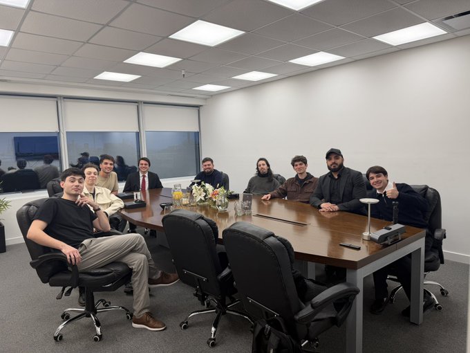 A group of people posing for a photo around a large wooden conference table in an office meeting room with overhead fluorescent lighting.