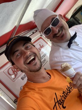 Two smiling men take a selfie under a tent; one wears an orange Airthreads shirt and holds an ice cream cone, while the other wears a white shirt, sunglasses, and a chef-style hat.