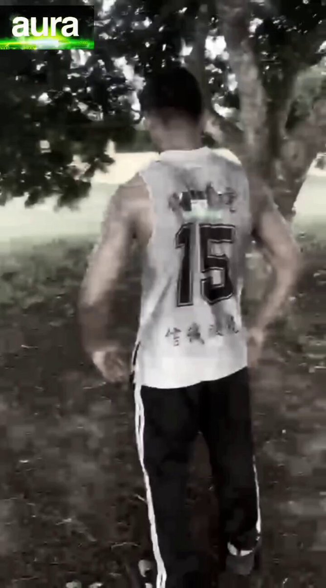 The image features a man wearing a gray tank top, black pants, and white shoes standing in front of a tree. He has a sticker on his back that reads " Aura" along with an accompanying caption about the aura community. The man is also holding a skateboard, which he appears to be using as part of his workout routine.
