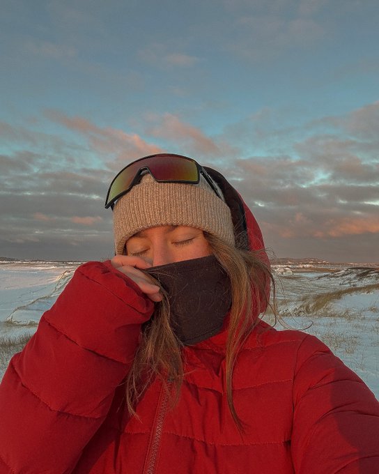 A selfie of a woman wearing a red puffer jacket, beige beanie with ski goggles, and a black face covering, standing in a snowy field at sunset.