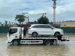 A photo of a white SUV being transported on a flatbed tow truck in a flooded area with construction cranes visible in the background.