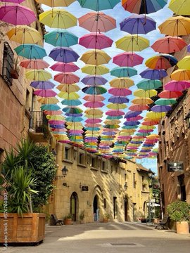A photograph of a narrow street with yellow and beige buildings, covered by a canopy of colorful umbrellas suspended in the air against a blue sky.