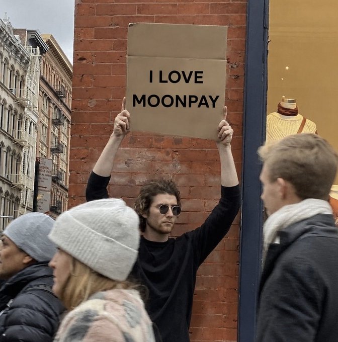 A photo of a man in sunglasses holding a cardboard sign that reads 'I LOVE MOONPAY' above his head while standing on a city street.