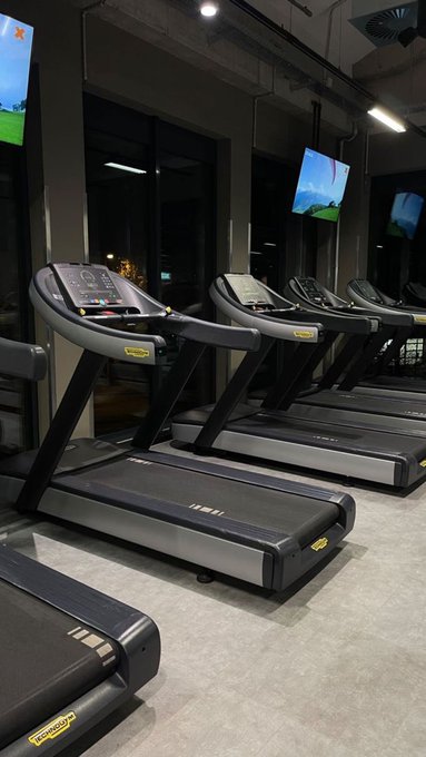 A row of Technogym treadmills lined up in a gym, featuring digital screens displaying a green landscape.