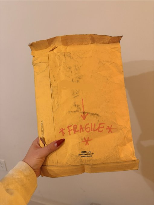 A hand holding a yellow padded mailer envelope marked with red handwritten '*FRAGILE*' text, arrows, and asterisks.