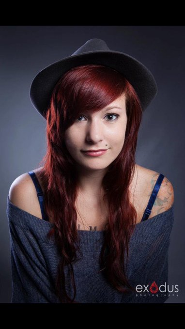 A portrait of a woman with long red hair wearing a black hat and a grey off-the-shoulder top, with the 'exodus premiere' logo in the corner.