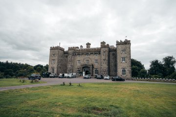 A large stone castle or manor house with multiple turrets and chimneys sits on a grassy lawn under an overcast sky, with several cars parked in front.