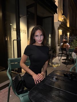 A woman in a black dress sits at an outdoor café table with a glass of white wine, captured in a moody evening setting under warm lighting. The caption "Hmmm" suggests a contemplative or skeptical mood as she appears lost in thought during her night out.