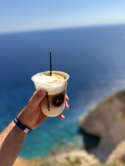 A hand holds a plastic cup of iced coffee labeled "Zakynthos" with a straw, set against a backdrop of a deep blue ocean and rocky cliffs.