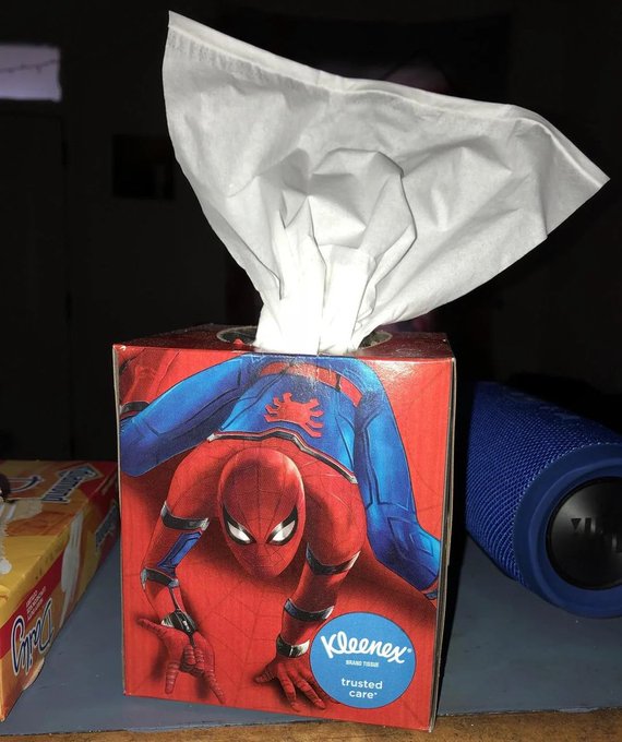 A box of Kleenex Brand Tissues featuring a Spider-Man design, labeled "trusted care," sits on a blue surface next to a blue Bluetooth speaker and a package of snacks.