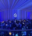 The image shows a large conference or event hall filled with attendees seated facing a stage illuminated with dramatic blue lighting and a prominent SUI logo displayed on the backdrop. This connects to the post "Gm $SUI agents," referencing the Sui blockchain ecosystem, suggesting the event is likely a Sui-related crypto conference or developer gathering.