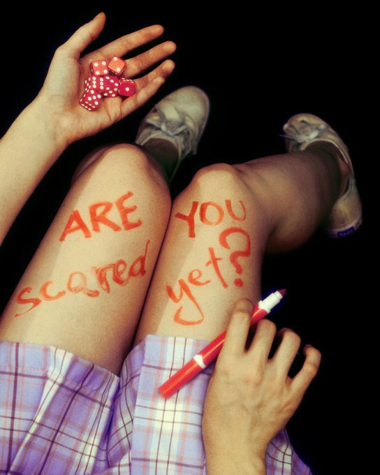 A first-person perspective photo looking down at legs with 'ARE you scared yet?' written in red marker on the skin, while one hand holds red dice and the other holds the marker.