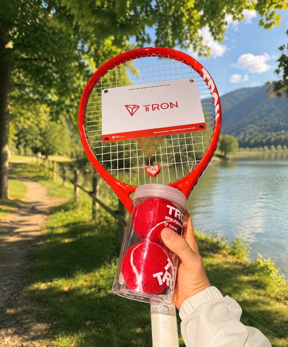 A hand holding a red Wilson tennis racket and a jar containing two red TRON-branded tennis balls by a lake with mountains in the background.