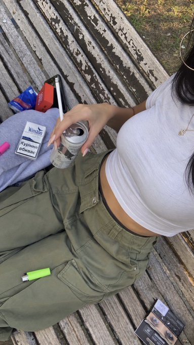 Overhead photo of a person sitting on a weathered wooden bench, wearing a white tank top and olive cargo pants, holding a can and a pen, with a Winston cigarette pack visible nearby.
