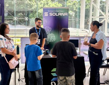 The image shows attendees at a Solana-branded booth during the Maker Fair at UCenfotec, where a representative engages with curious students and entrepreneurs interested in building on the Solana blockchain.

Three people interact at a booth featuring a Solana banner ("The Internet Capital Solana"), with two young boys in casual clothing asking questions while a man and woman converse about the platform, illustrating the post's message about engaging Costa Rica's next generation of developers with the Solana ecosystem.