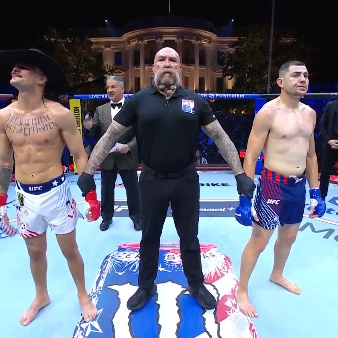 The image features a group of men standing around each other, with one man wearing a black shirt and holding a boxing glove while another man stands next to him. The third man has his arms crossed over his chest. They are all dressed in shorts and appear to be at a UFC event or competition. In the background, there is an official who seems to have secured the victory for the fighter with the TKO.