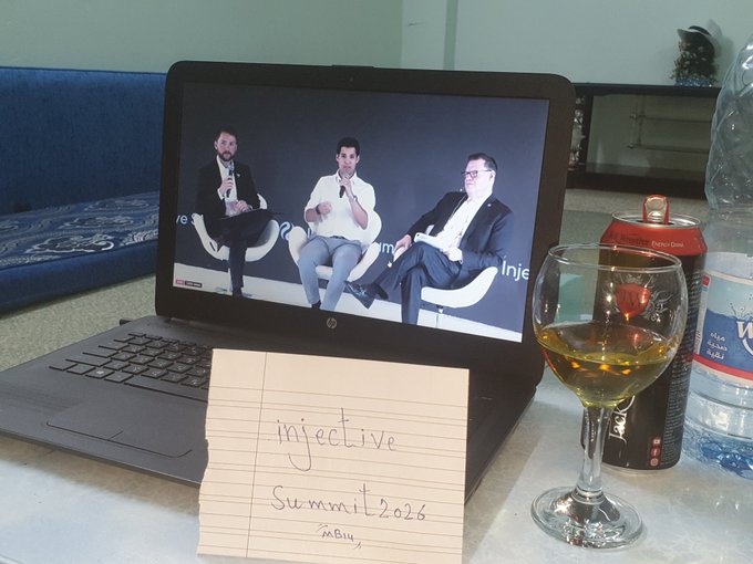 A laptop on a table showing a live stream of three men in suits at a panel discussion, with a handwritten note reading 'injective summit 2026 in Bkk', a glass of white wine, a soda can, and a water bottle nearby.