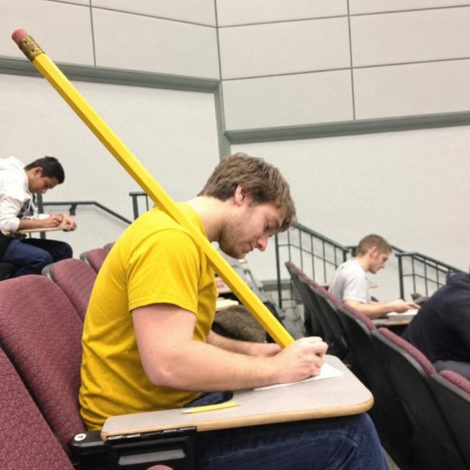 A student in a yellow shirt sits at a tiered lecture hall desk, intensely focused on taking notes with a pencil, while a comically oversized yellow pencil with a pink eraser is balanced on his shoulder—visually exaggerating the act of "noting" something down to humorous effect in response to a simple "noted" reply.
