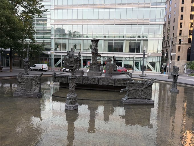An ornate fountain with sculptural figures sits in a reflecting pool in front of a modern glass building in an urban plaza.
