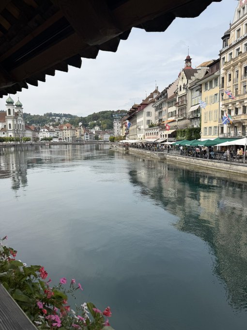 A picturesque view of a European town along a calm river, with historic buildings, outdoor cafés, and a church tower reflected in the water—the kind of serene, postcard-perfect setting that makes the good times feel even sweeter. The image pairs with the post by evoking a sense of enjoying life's beauty while the markets are thriving.