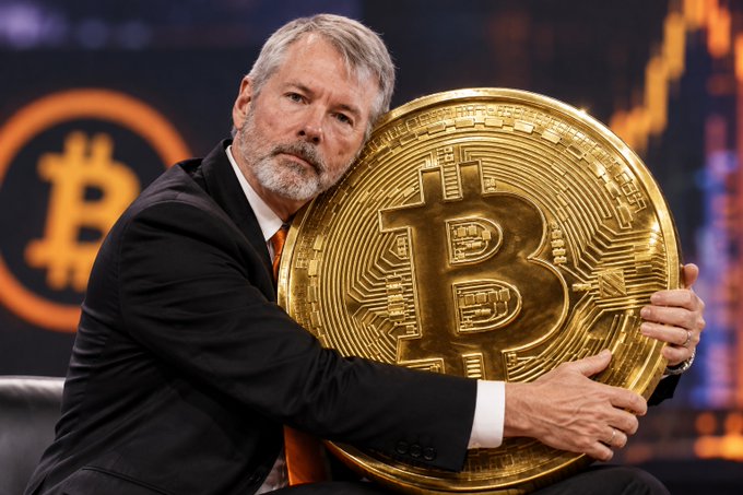 Michael Saylor, known for his Bitcoin advocacy, holds a large physical Bitcoin token while wearing a suit in front of a backdrop featuring the Bitcoin logo.