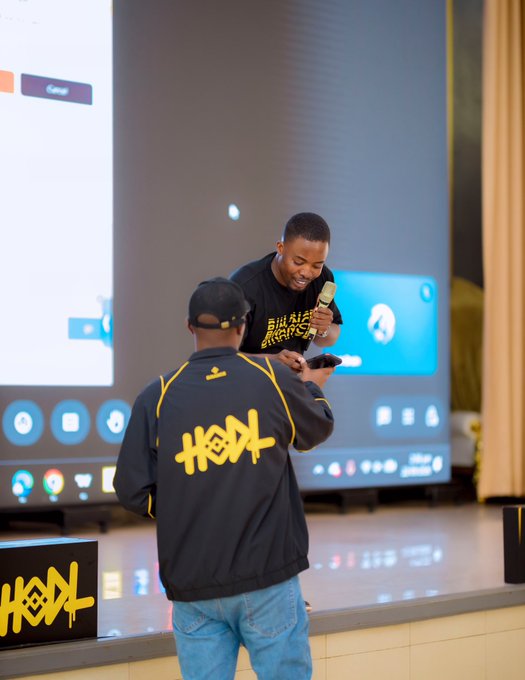 Two men at an event wearing matching black jackets with yellow 'HODL' lettering; one holds a microphone while the other looks at a phone, with a large screen behind them.