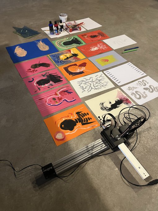 An overhead view of an art workspace with various colorful paintings, stencils, and ink bottles spread across a concrete floor next to a tripod stand.