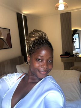 A woman with braided hair in a high bun poses for a selfie in a hotel room, wearing a light blue satin top and smiling warmly at the camera. The casual setting, with a bed and warm lighting in the background, fits the relaxed vibe of capturing a personal moment at the Solana summit.