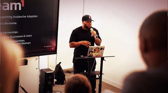 The image shows a man standing at a podium, speaking into a microphone while holding a laptop computer. He appears to be giving a presentation or lecture. The background of the image features a white wall with a banner that reads "Built for Business @avax @AvaxTeam1". This suggests that the event might be related to business or technology-related topics and is being held in an office setting.