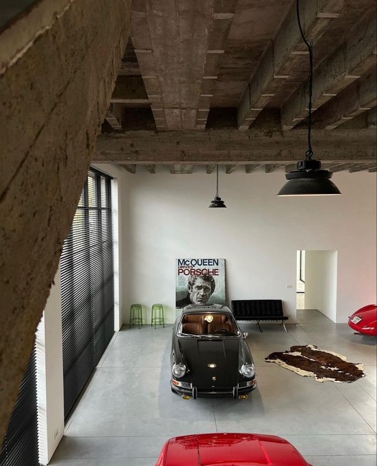 A spacious garage with exposed concrete beams features a black classic Porsche, a 'McQueen Porsche' poster, a red car in the foreground, and modern black pendant lighting.