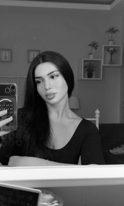 A black-and-white mirror selfie of a young woman with long dark hair, wearing a scoop-neck shirt and a delicate bracelet, posing with her phone as she captures the shot. The "Gm Sunday" caption pairs with her relaxed Sunday morning vibe, enhanced by the cozy bedroom setting with decorative shelves and a small framed sign in the foreground.
