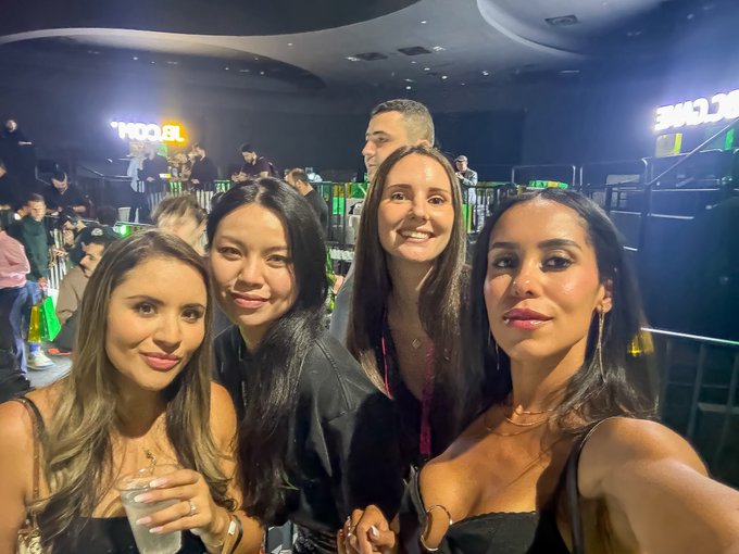 Four women smiling for a group selfie in a dark venue, with a 'WCDUB' neon sign and other patrons visible in the background.