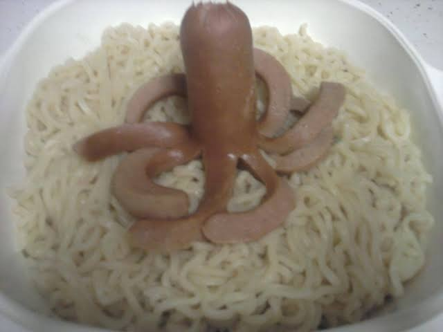 A bowl of noodles topped with sausage pieces arranged in an unusual decorative pattern resembling a flower or symbol.