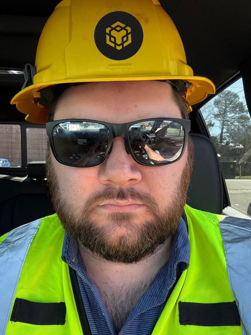 A construction worker in safety gear taking a selfie in his vehicle. The yellow hard hat features a black circular logo with a cube design, referencing the BNB Chain branding in the caption about "building" on the blockchain.