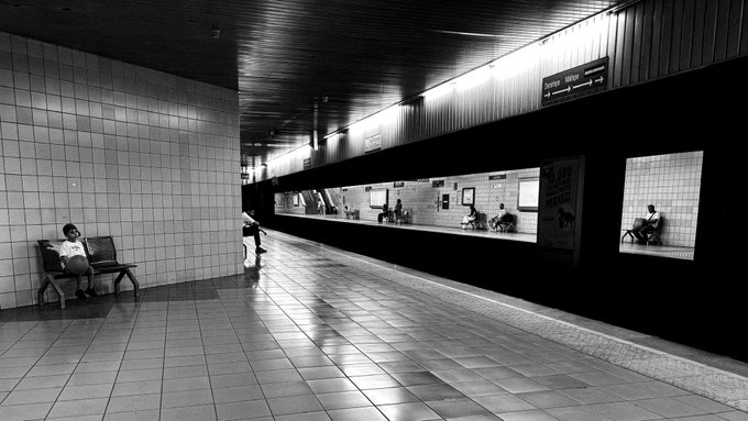 A black-and-white photo of a subway station with a person sitting on a bench on the left, while a train is visible on the right with passengers inside.