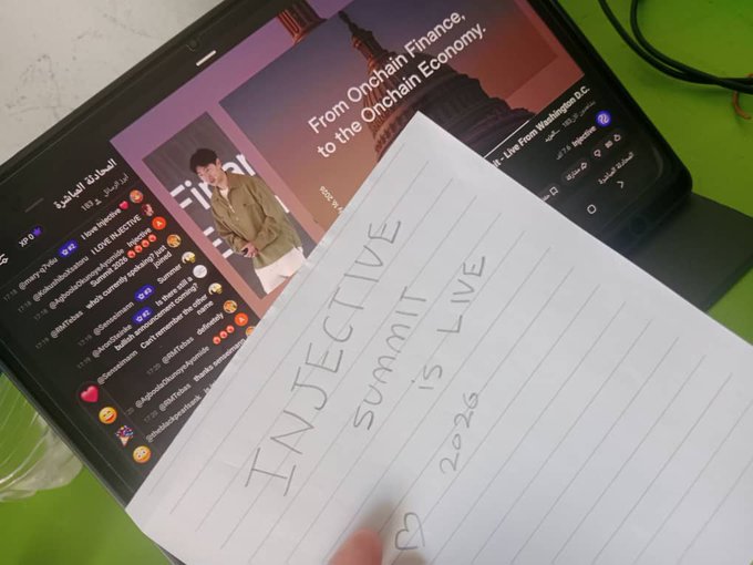 A handwritten note reading 'INJECTIVE SUMMIT IS LIVE 2026' rests over a tablet displaying a live stream titled 'From Onchain Finance to the Onchain Economy' with Arabic chat messages visible.