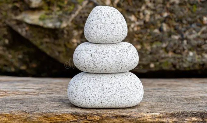 The image features a stack of three white rocks or pebbles arranged in a pyramid shape on a wooden surface. The rocks are positioned at different heights, creating an interesting visual effect. The background appears to be a blurred outdoor scene with trees visible through the foliage.