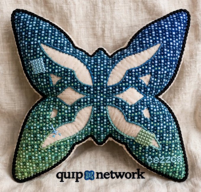 A butterfly-shaped cross-stitch pillow in shades of blue, green, and cream rests on a linen background, branded with the 'quirp network' logo and watermark 'BeZZa'.