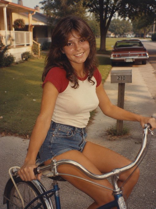 A vintage-style photo of a young woman with brown hair wearing a red and white raglan t-shirt and denim shorts, sitting on a blue bicycle on a suburban street with a mailbox displaying '210'.