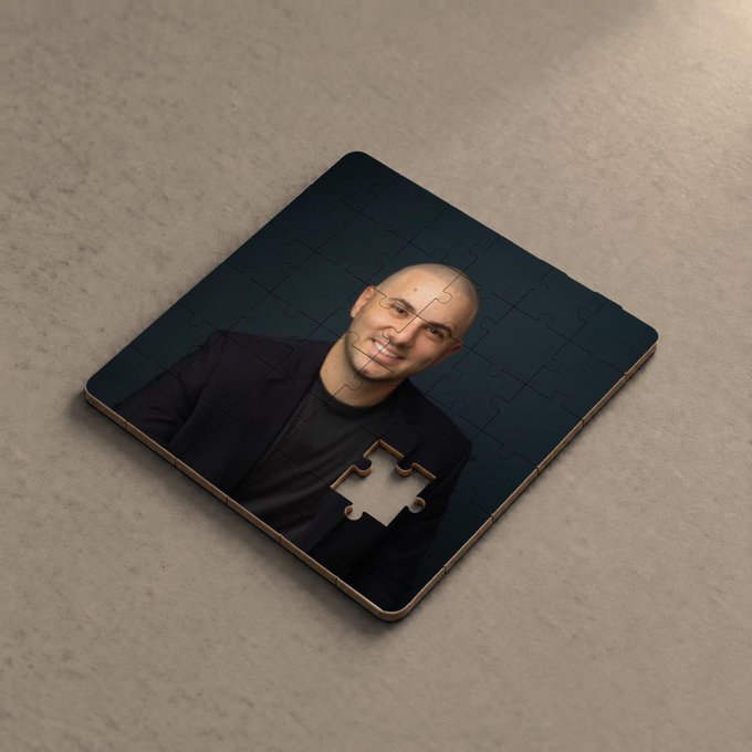A nearly completed jigsaw puzzle resting on a concrete surface depicts a portrait of a smiling bald man in a black suit, with one missing piece visible in the lower center.