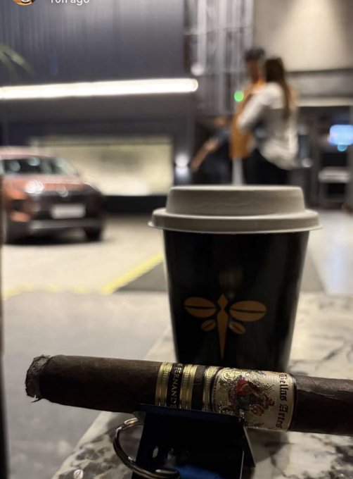 A cigar labeled 'Trinidad Fundadores' rests on an ashtray next to a black coffee cup, with a car visible in the background of what appears to be a parking garage.