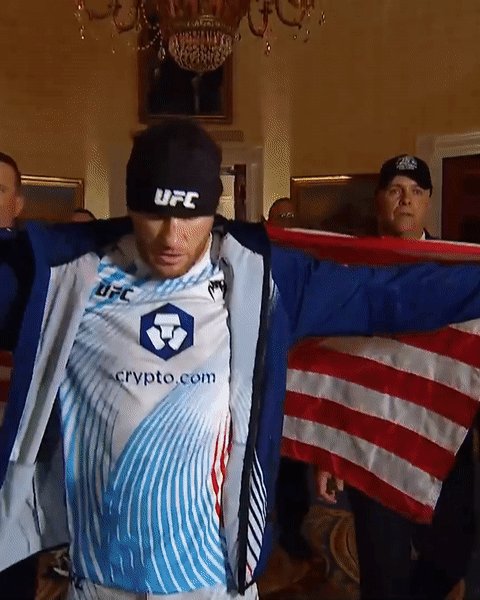 The image features a man wearing an UFC jersey, standing next to a flag with red, white, and blue stripes. The man appears to be adjusting his jacket or putting something on his face while standing near the flag. In the background, there are other people present who seem to be watching the scene. The post includes text that reads "Making the walk to the Octagon with @Justin_Gaethje is NYC First Responder: Thomas O'Connor & Medal of Honor Recipient: Lance Corporal Kyle Carpenter". This suggests that the man in the UFC jersey might have received an award or recognition, possibly for his performance in a fight.